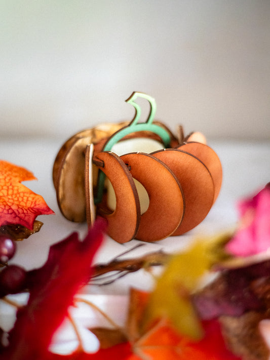 Miniature 3D wooden pumpkin decoration with orange painted panels and green stem surrounded by colorful autumn leaves