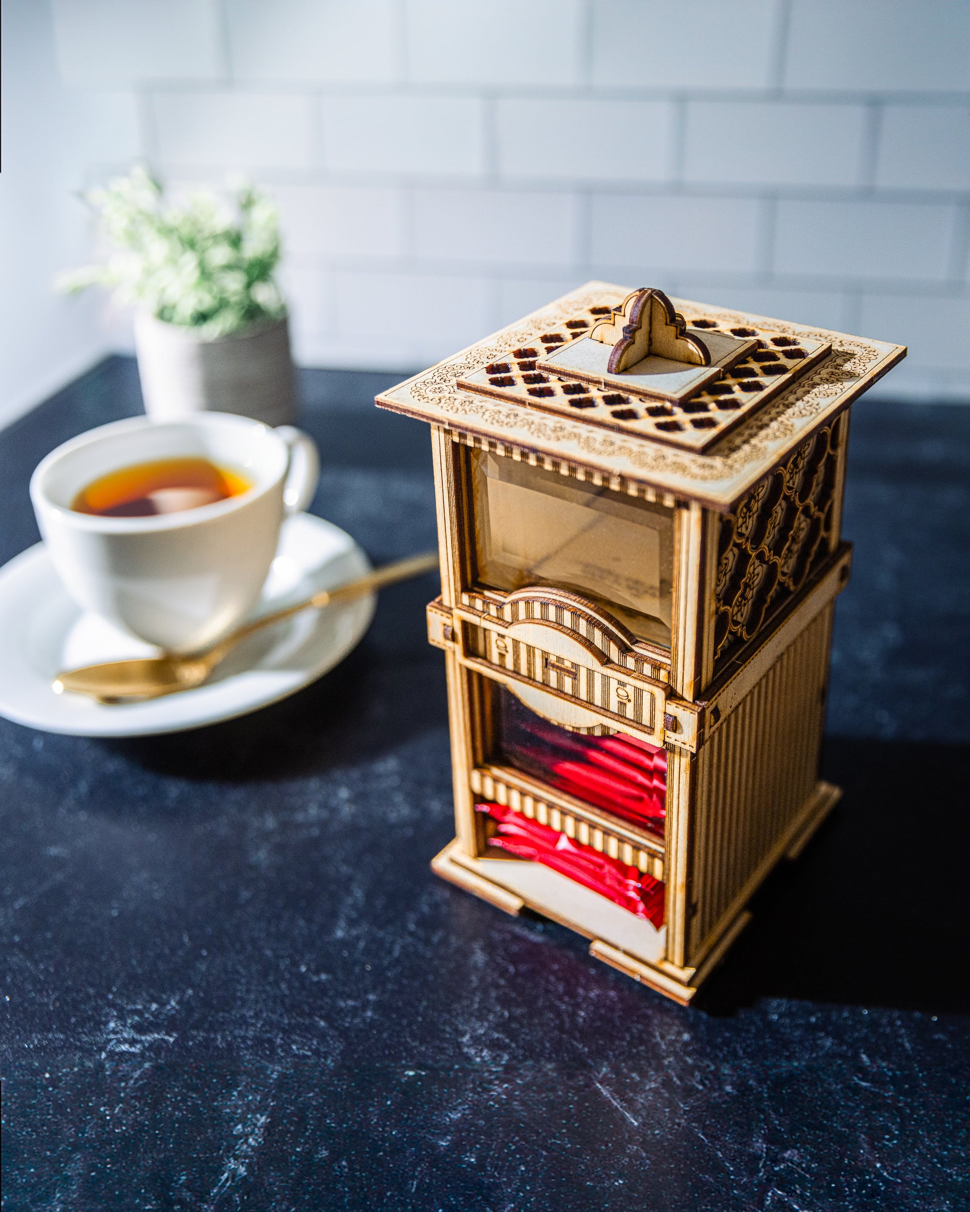 A wooden 3D kit assembled into a tea bag dispenser and cookie jar combination, with a cup of tea in the background.