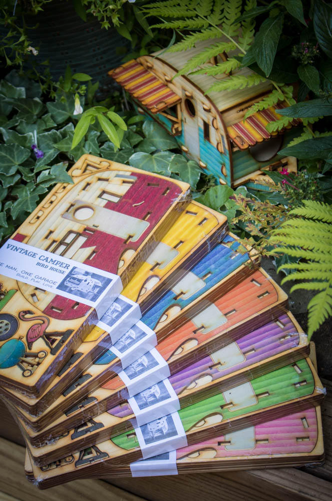 stacked display of camper birdhouse color options showing variety of painted finishes in their flat packaging. Red, yellow, blue, orange, purple, green, and pink.
