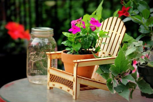 A miniature Adirondack chair made of birch wood, designed to be used as a plant stand or drink holder, placed next to a pot with a pink flower in it.