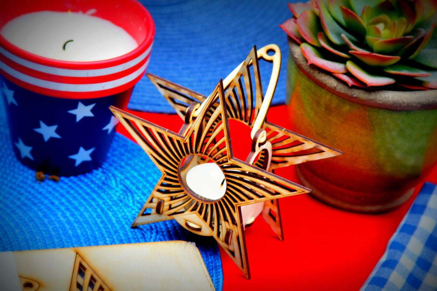 A hanging tealight luminaire kit designed as a star-shaped wooden lantern with a visible hanging hook, displayed on a table with a red and blue background, alongside a cups and a potted plant.